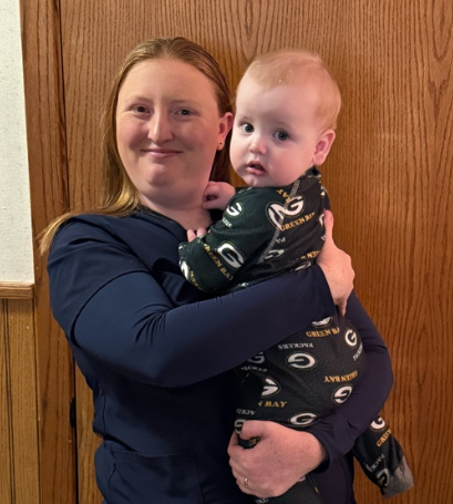 Melanie Ungerecht, founder of Melanie’s Moon Beans, in dark blue scrubs, smiling as she holds her baby, Brett, who is wearing Green Bay Packers pajamas.