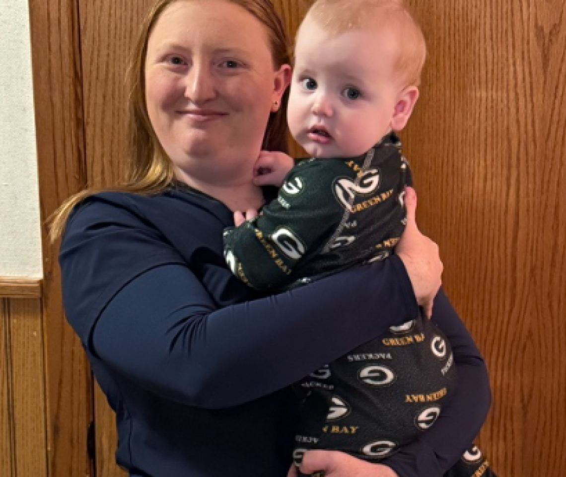 Melanie Ungerecht, founder of Melanie’s Moon Beans, in dark blue scrubs, smiling as she holds her baby, Brett, who is wearing Green Bay Packers pajamas.
